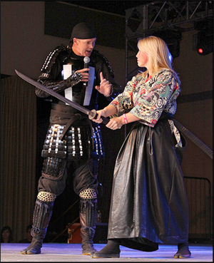 Samurai Dan is a tall man in a skull cap and black samurai-style armor.  He holds a microphone and holds his other hand demonstratively in front of Jillian who wears kimono and hakama and holds a katana.