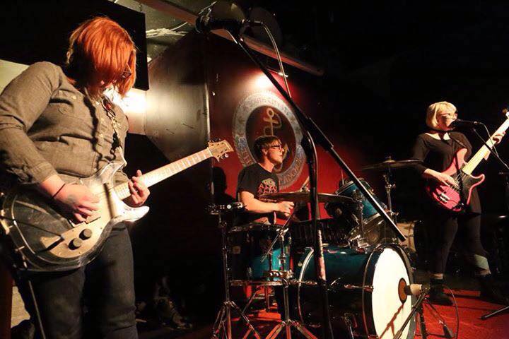 a redhead rocks out in the foreground while her other band members look fierce on a small bar stage